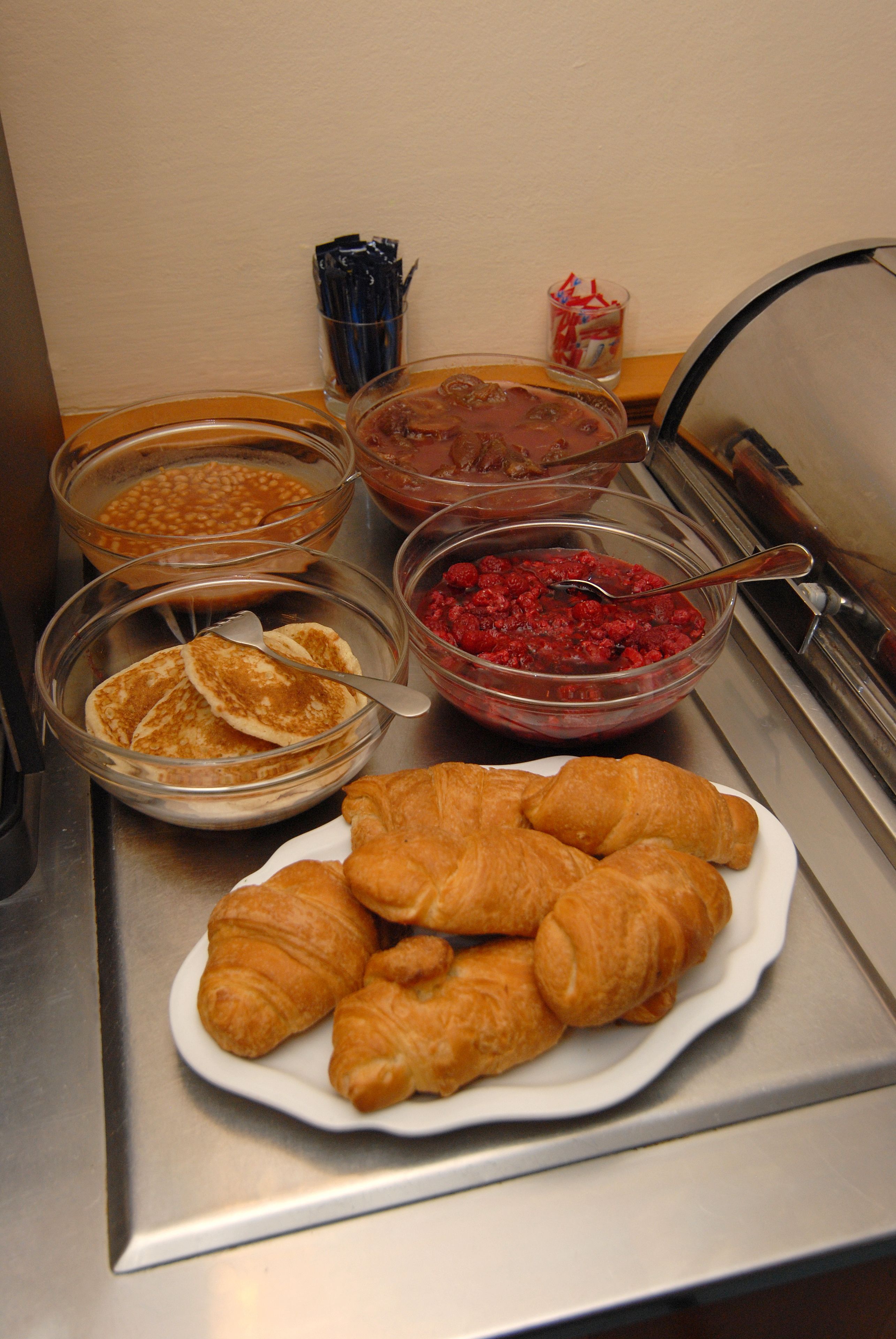 A buffet with various food items including pancakes, a bowl of red fruit compote, and croissants on a plate.