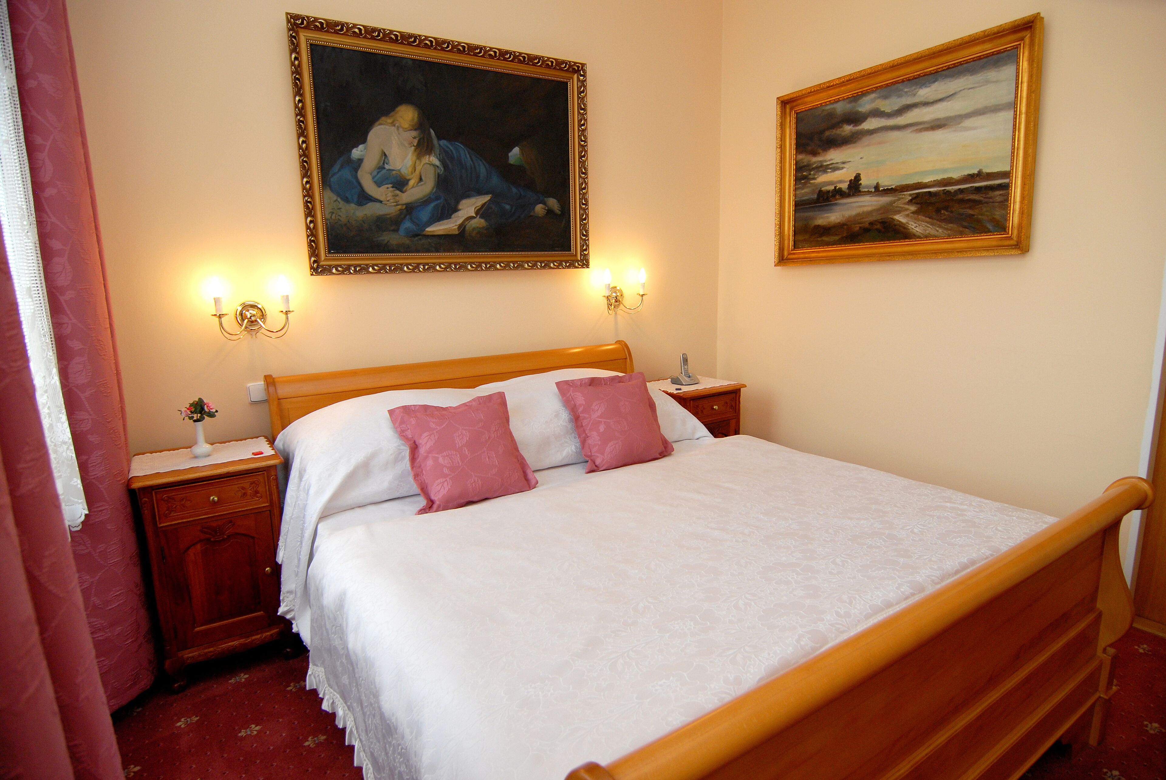 A neatly made bed with a wooden frame, two bedside tables with lamps, and two paintings on the wall.