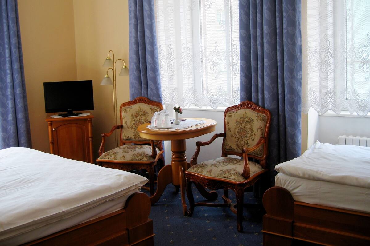 Quarto de hotel com duas camas de solteiro, uma mesinha com um vaso e um livro, duas cadeiras, uma televisão sobre um suporte e uma janela com cortinas.