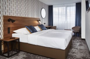 Una camera d'albergo con un letto grande, una testiera in legno, un comodino con una lampada, una piccola scrivania e una sedia.