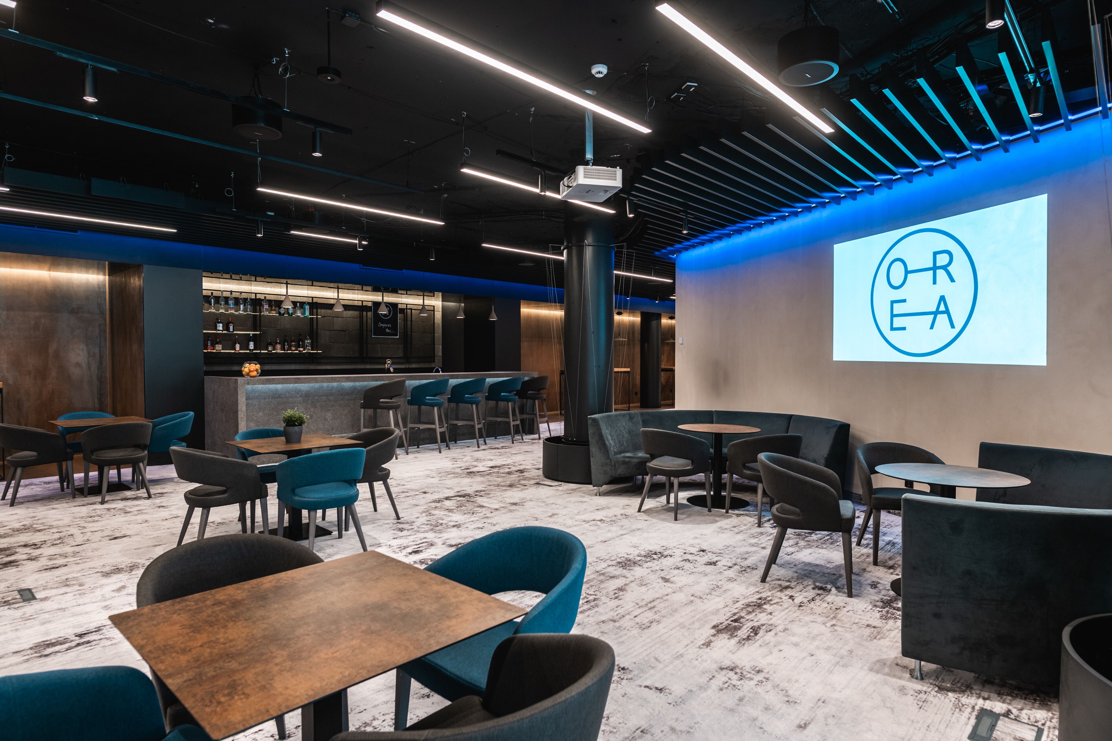 A modern interior with a bar area, seating arrangements, and a projection screen displaying "OREA".