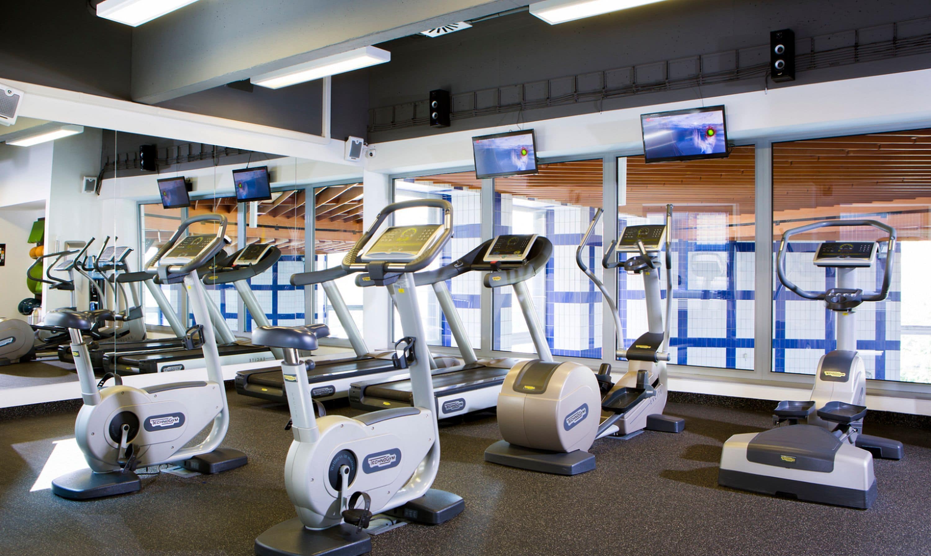 Un gimnasio con varias cintas de correr y elípticas, ventanas amplias con persianas y una pared de azulejos.