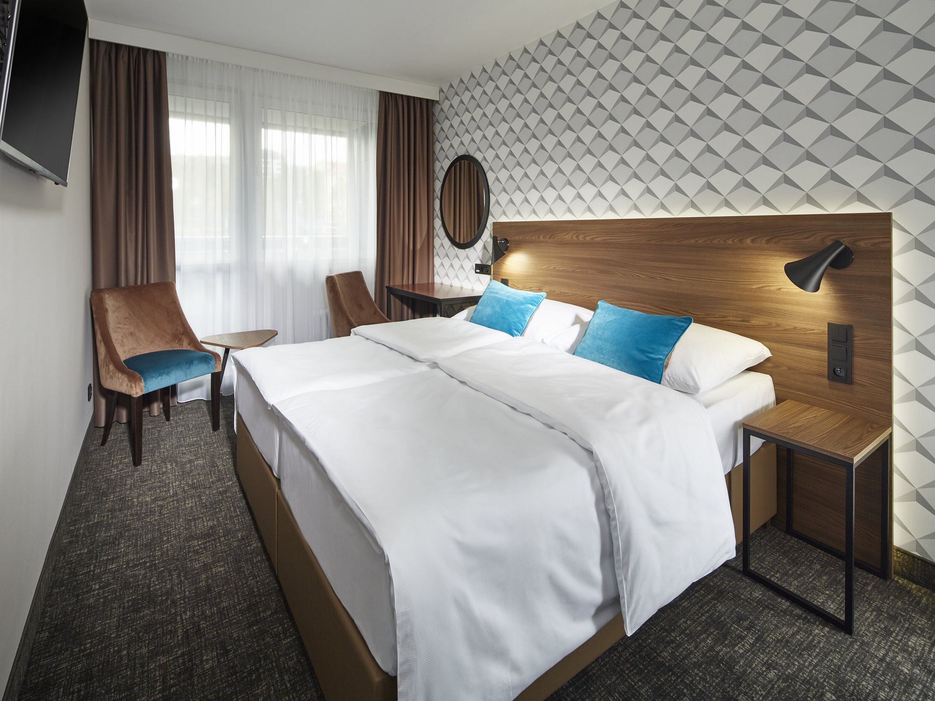 Ett hotellrum med en stor säng, ett skrivbord, en stol, en tv och en vägg med ett geometriskt mönster.