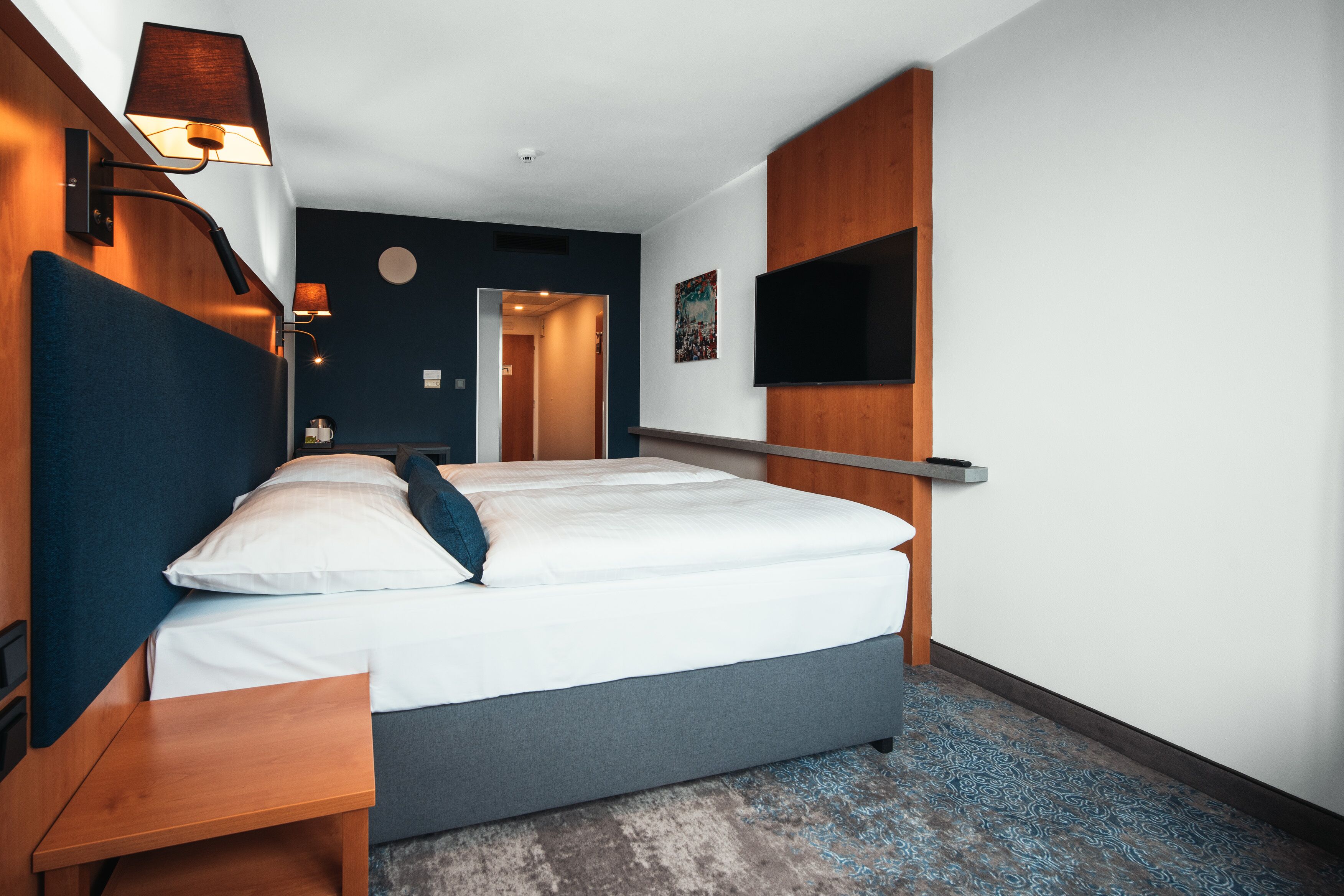 Una habitación de hotel moderna con una cama grande, mesitas de noche, un televisor de pantalla plana montado en la pared y un cabecero de madera.