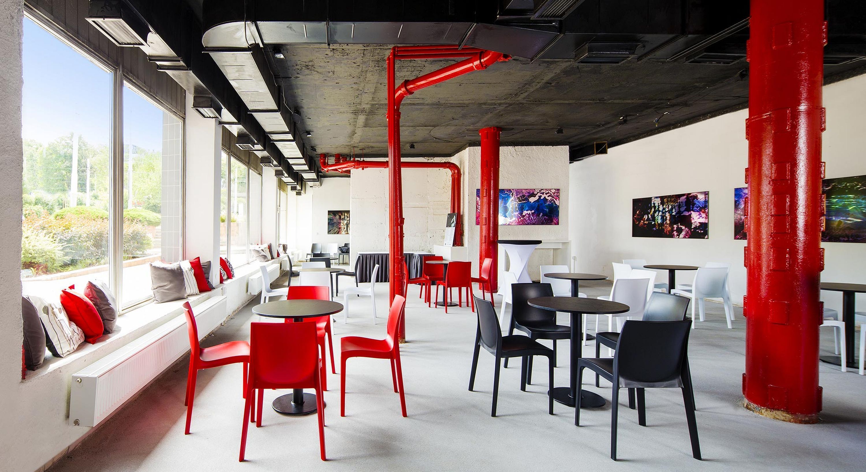 Un caffè moderno con sedie rosse e nere, tavoli bianchi e ampie vetrate sulla vista del verde.