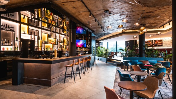 Zona de bar con mostrador, taburetes y estantes llenos de botellas. Hay mesas y sillas en la zona de estar.