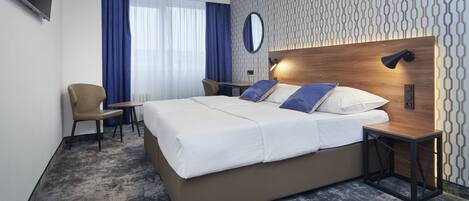 A modern hotel room with a large bed, a desk, a chair, and a TV mounted on the wall.