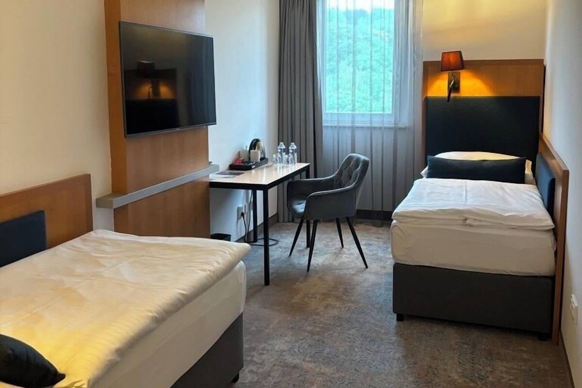 A hotel room with two beds, a desk, a chair, a TV, and a window with curtains.