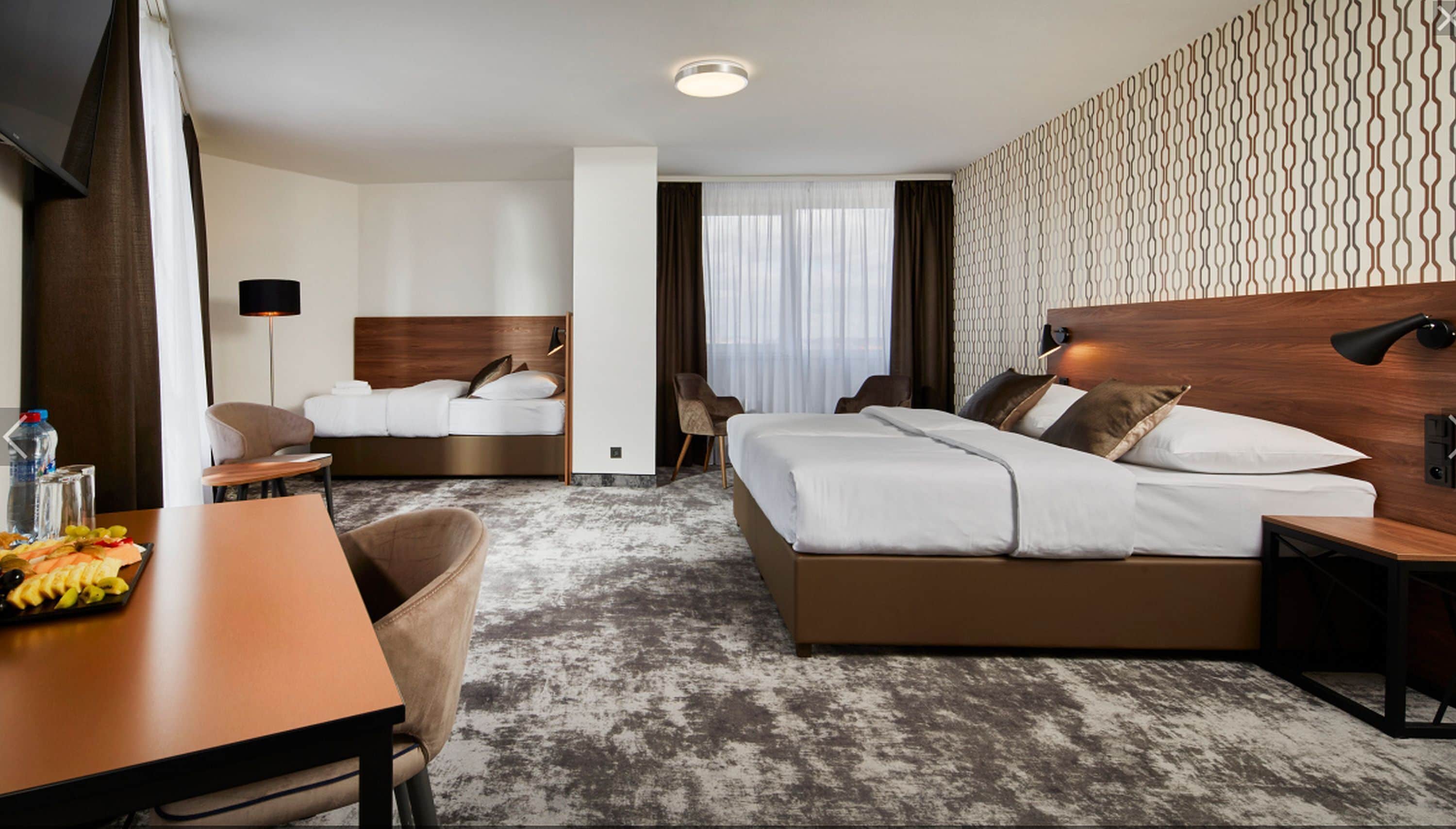 A hotel room with a large bed, a smaller bed, a desk with a chair, a table with fruit, and a patterned wall.