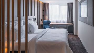 A modern hotel room with a large bed, a desk, a chair, and a window with curtains.