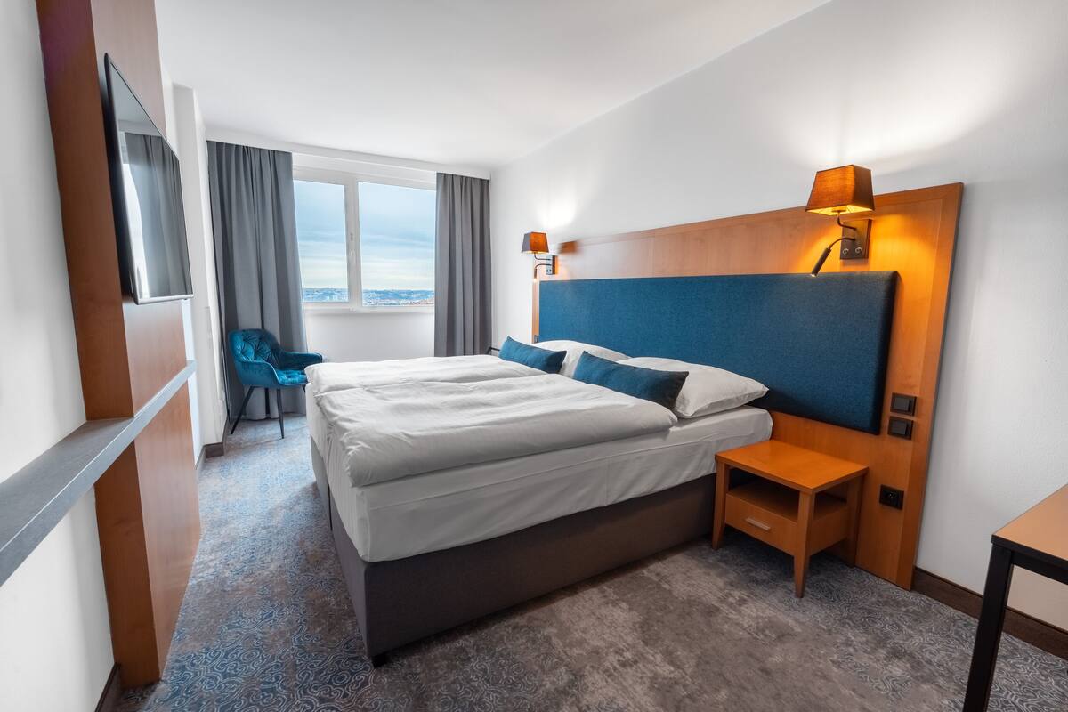 A hotel room with a large bed, a TV mounted on the wall, a small bedside table, and a view of the city through the window.
