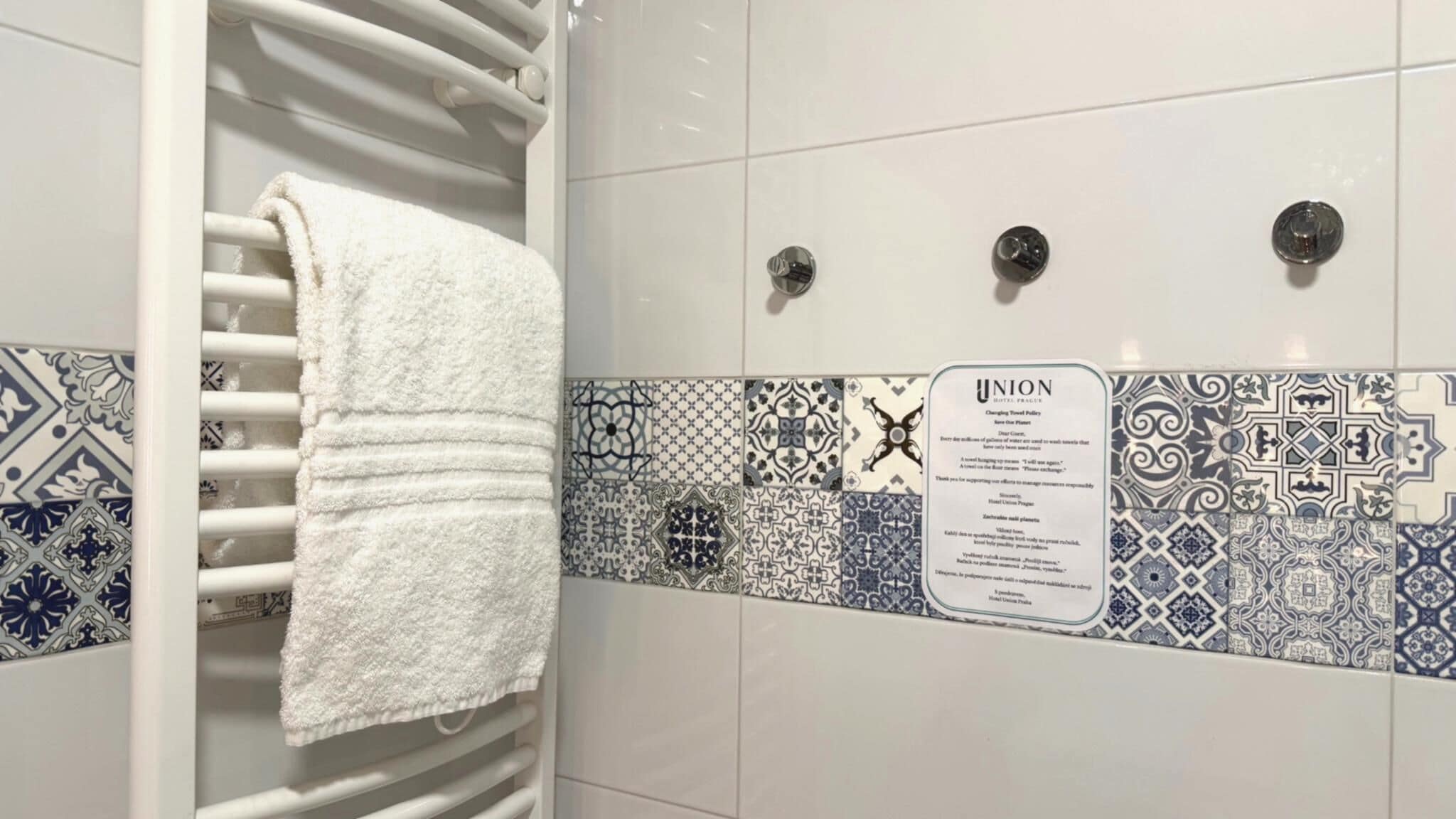 Un baño con una toalla blanca en un perchero, baldosas estampadas y un cartel sobre la política de toallas del hotel.
