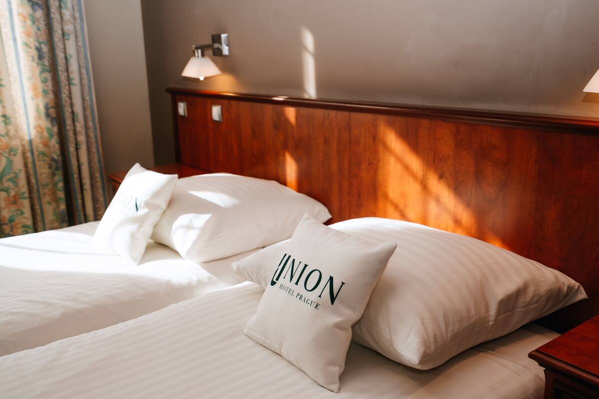 Quarto de hotel com duas camas, um painel de cabeceira de madeira e um travesseiro com a inscrição "UNION HOTEL PRAGUE".