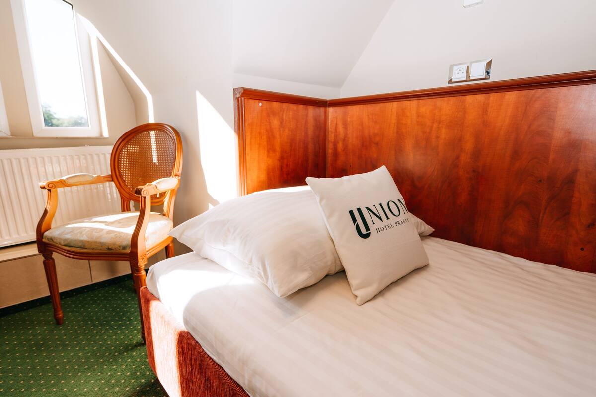Quarto de hotel com cabeceira de madeira, cama com roupa de cama branca, tapete verde e uma cadeira de madeira.