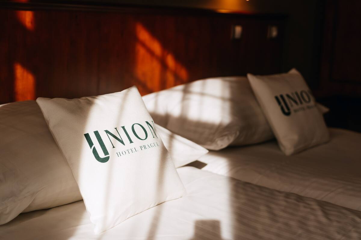 Uma cama com almofadas identificadas com "UNION HOTEL PRAGUE" e uma cabeceira de madeira.