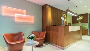 Un área de recepción con un mostrador de madera, dos sillones de cuero, una mesita con un jarrón de flores e iluminación moderna en la pared.