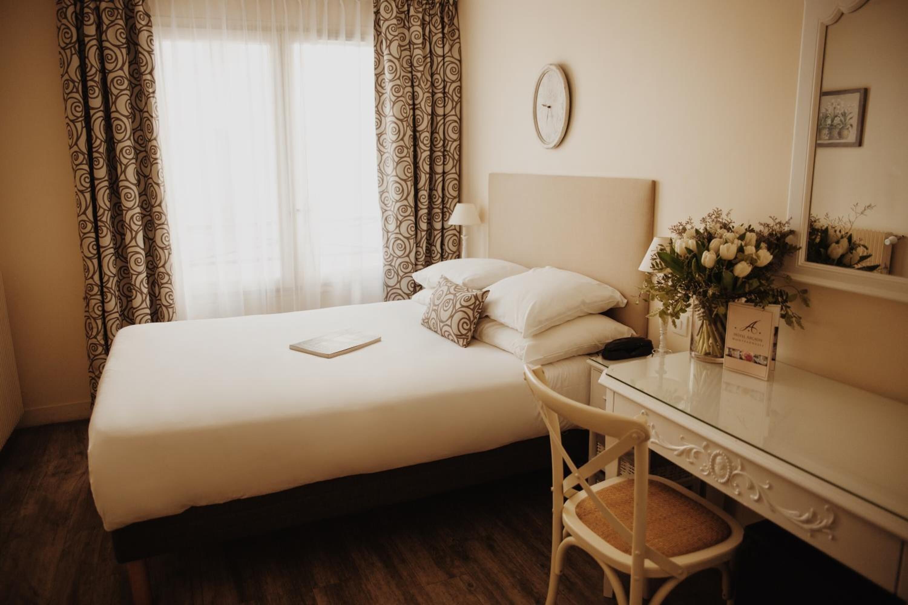 Habitació d'hotel amb un llit, una taula de treball, una cadira, una làmpada, un rellotge i un ram de flors.