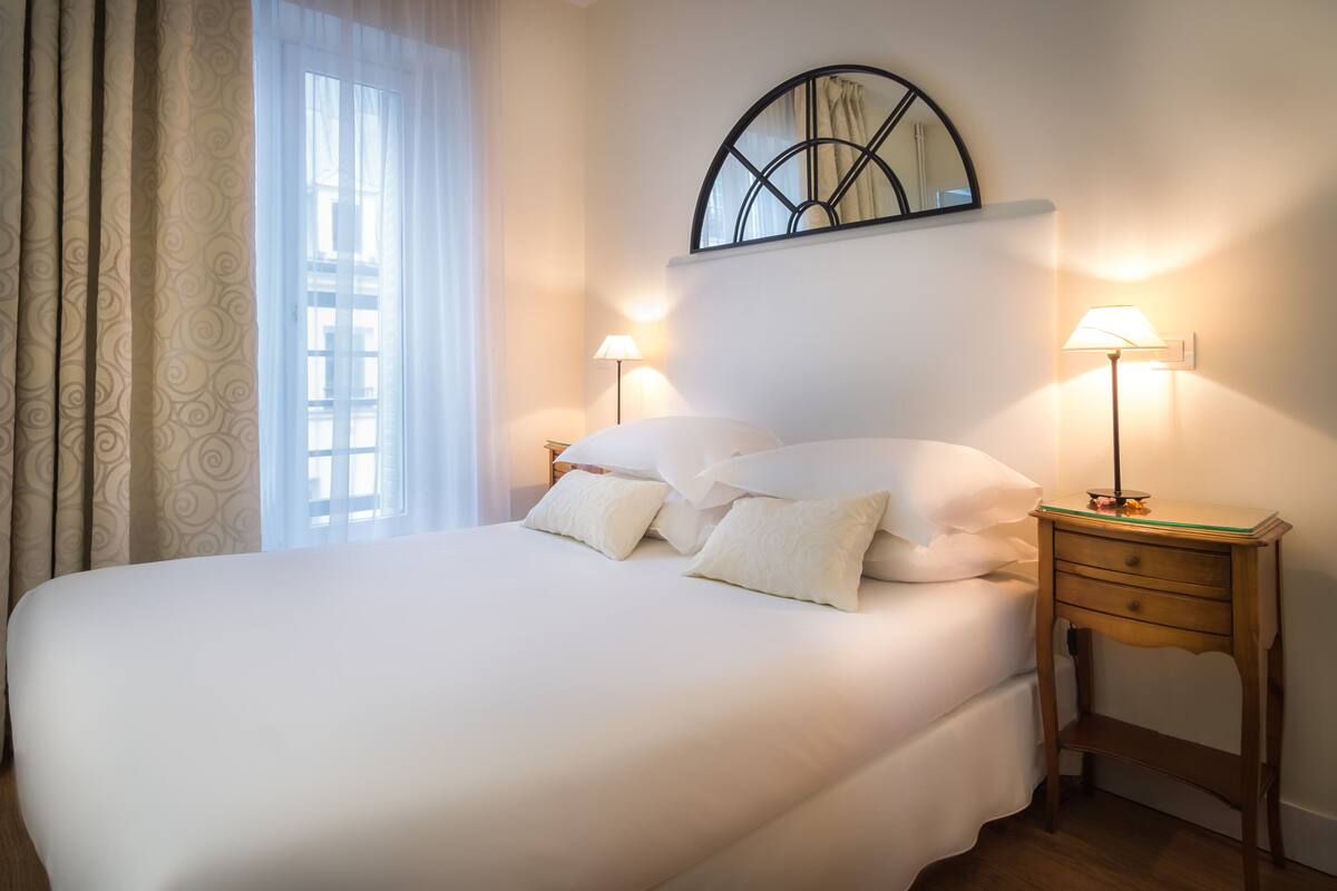 Un letto rifatto con lenzuola e cuscini bianchi, un comodino con una lampada e una finestra con tende.