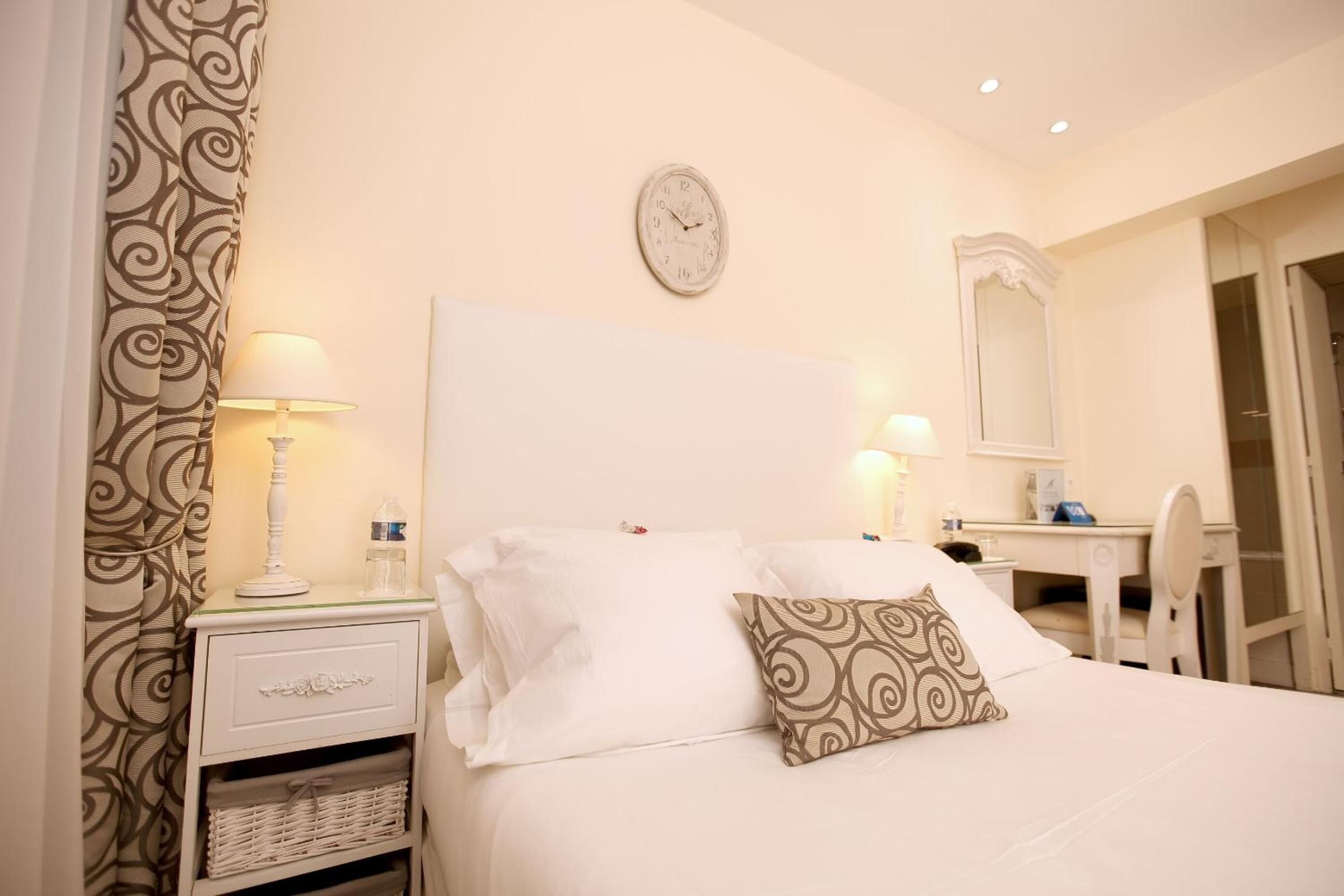A neatly arranged hotel room with a bed, bedside table, lamp, and a clock on the wall.