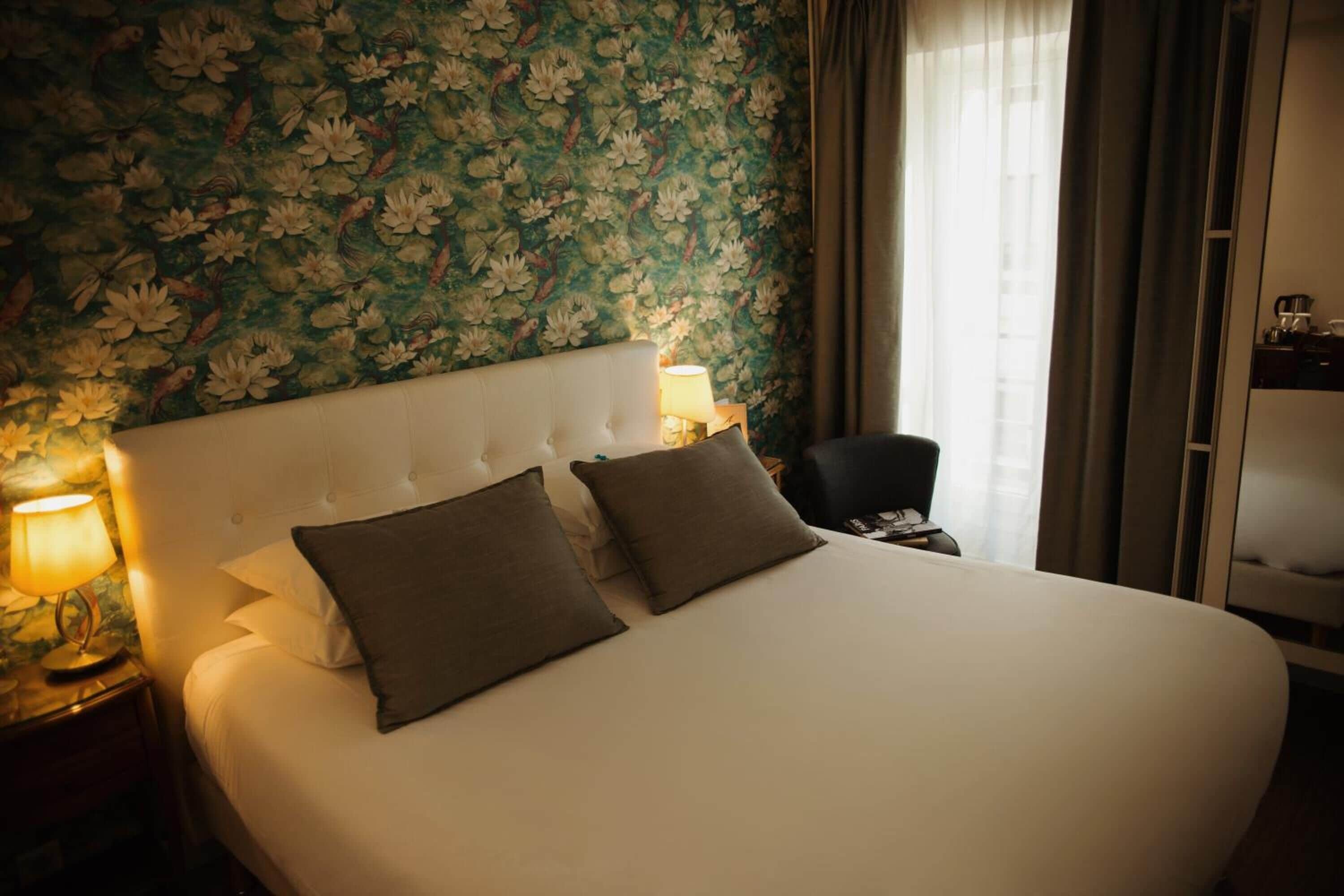 A hotel room with a large bed, two pillows, a nightstand with a lamp, and a floral wallpaper.