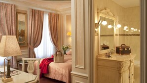 A room with a bed, a desk with a lamp, a mirror, and a bathroom with a sink and lights. - Hôtel & Spa Saint Jacques (Paris)