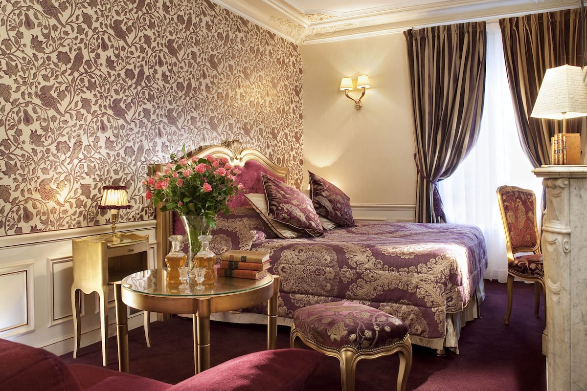 A luxurious bedroom with a large bed, ornate wallpaper, a round table with a lamp, and a chair.
