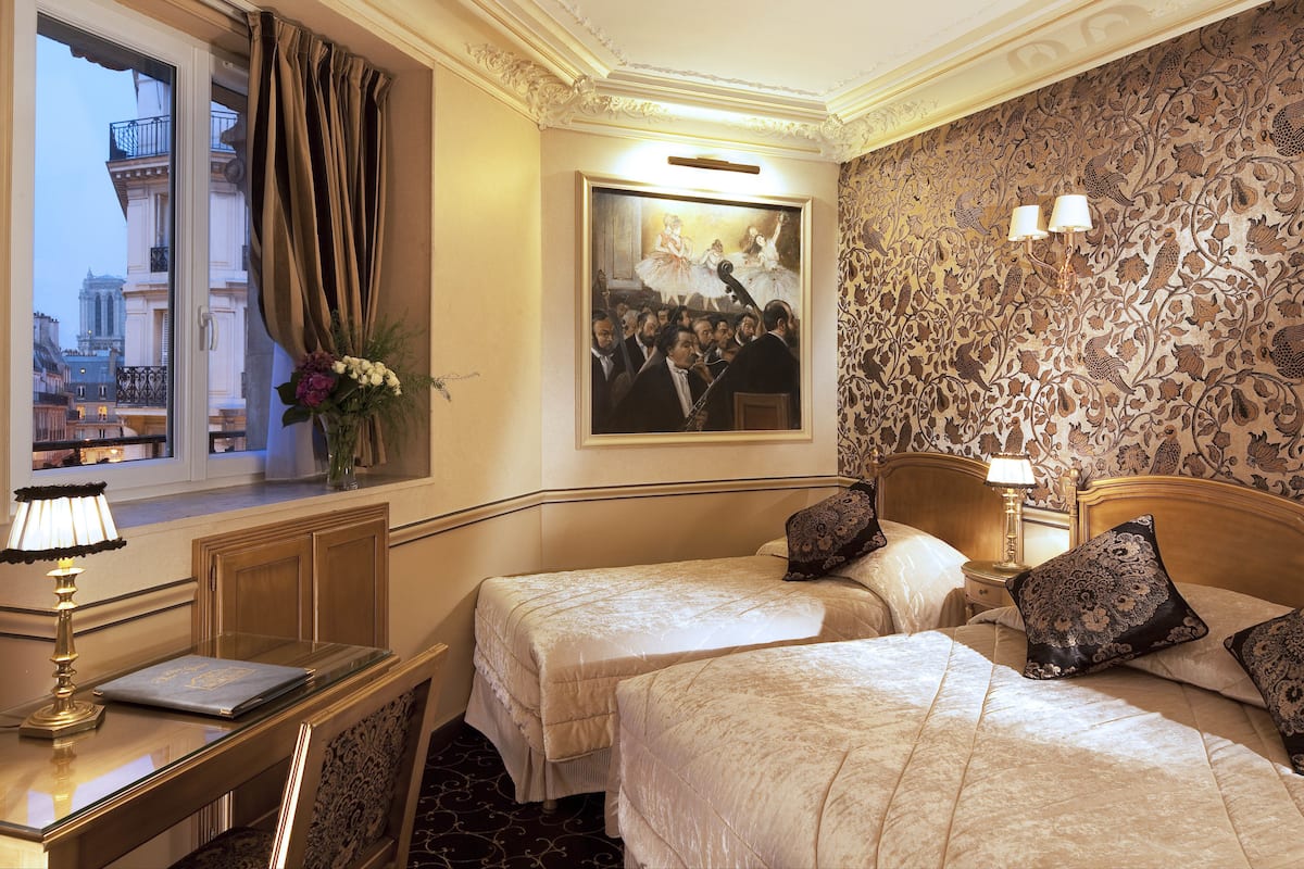 A hotel room with two beds, a desk with a lamp, a painting on the wall, and a view of buildings through a window.
