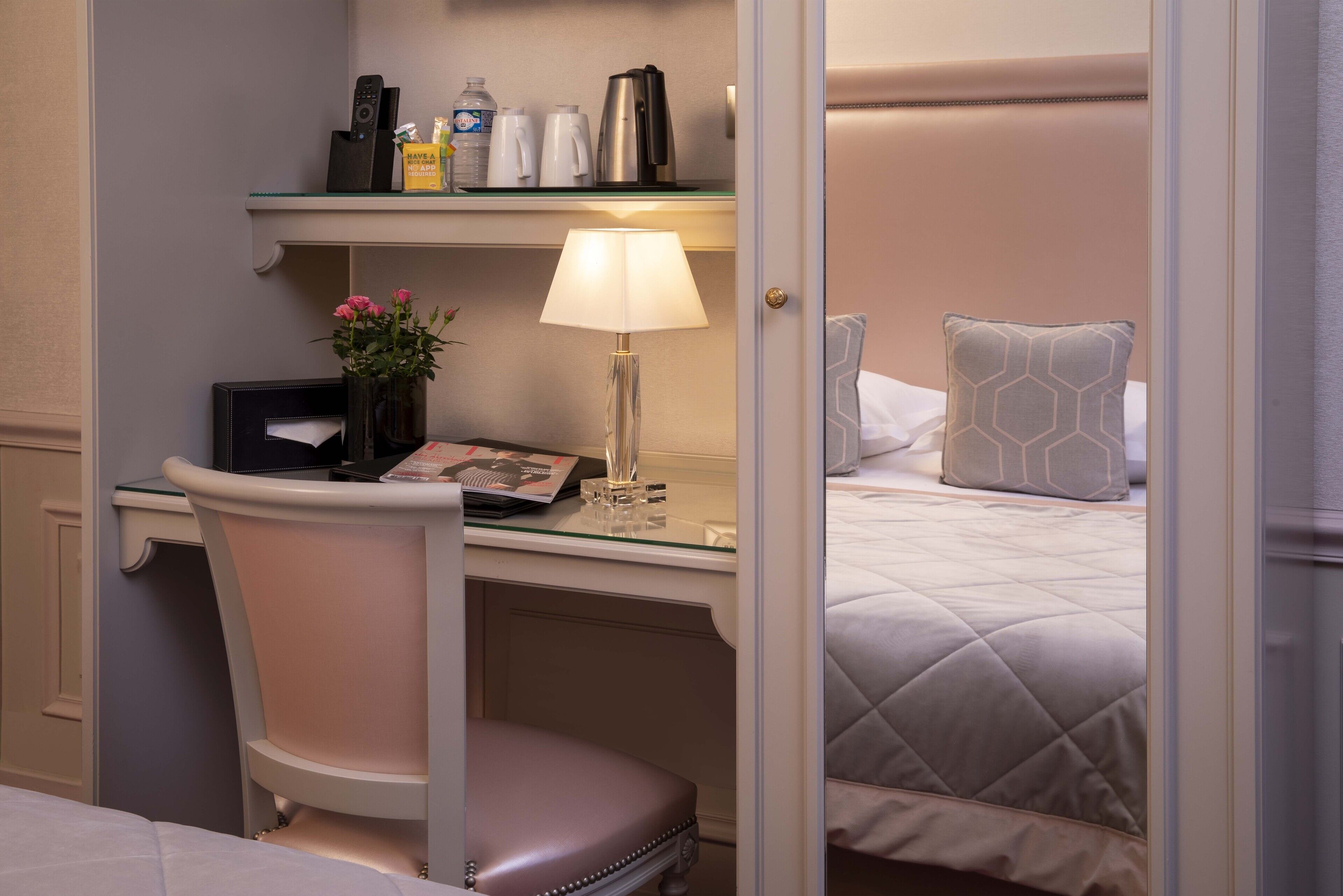 A hotel room with a bed, a desk with a lamp, a chair, and a shelf with various items.