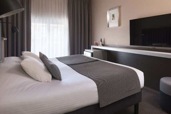 A modern hotel room with a large bed, a flat-screen TV mounted on the wall, a desk with a chair, and a window with curtains. - Hotel Diana Dauphine (Strasbourg)