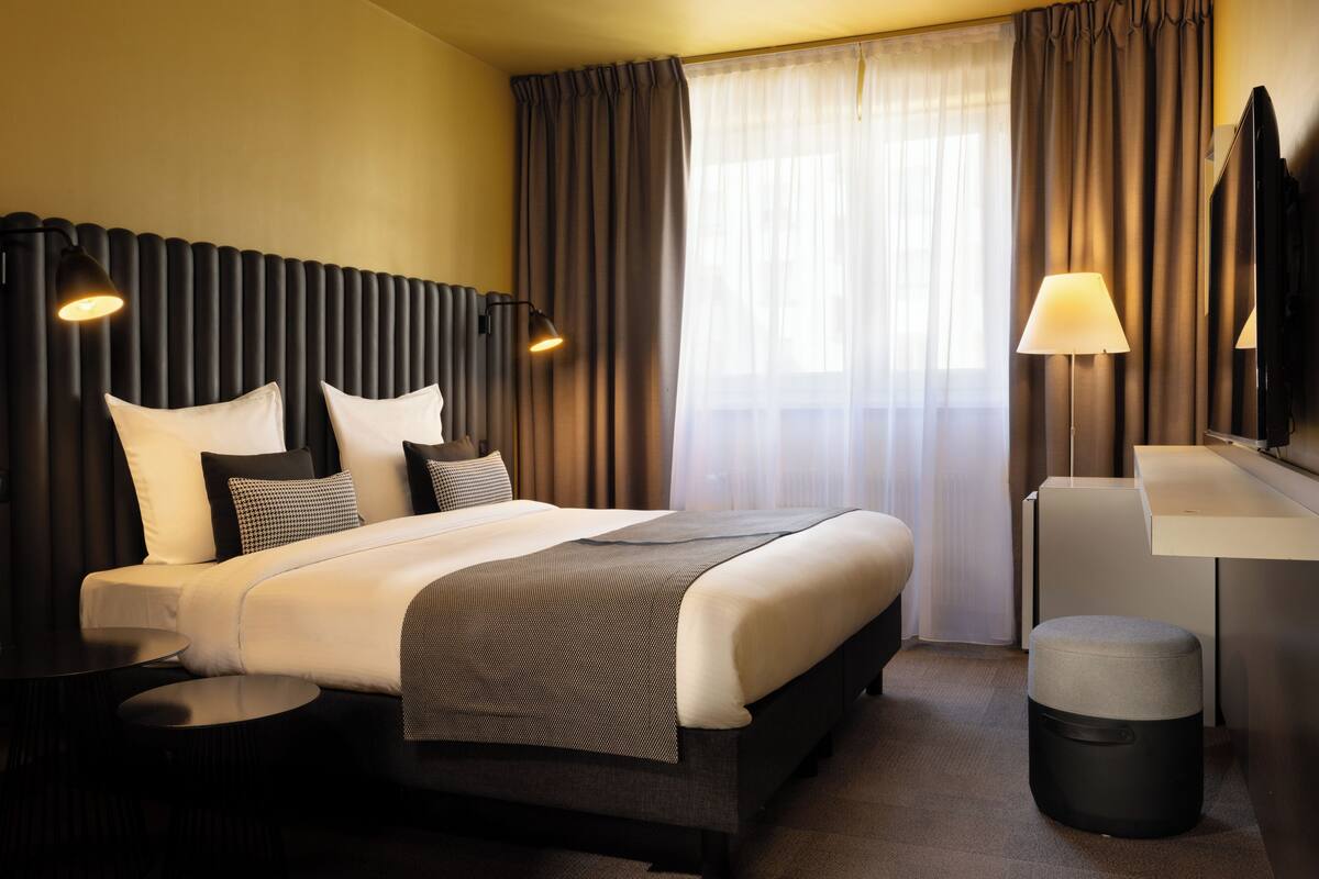 Une chambre d’hôtel moderne équipée d’un grand lit, de deux tables de chevet, d’un bureau, d’une chaise, d’un miroir et d’une poubelle.