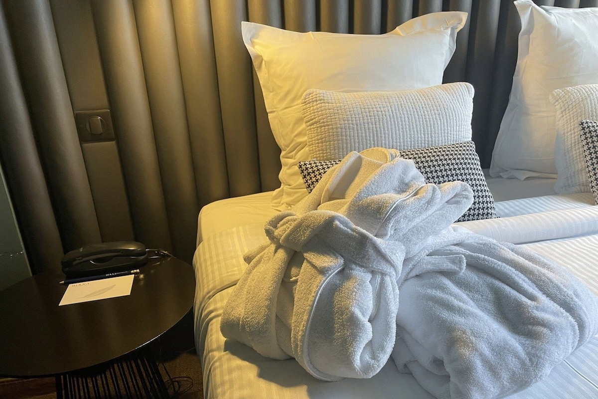 A neatly made bed with white bedding, a pair of white bathrobes, and a black side table with a mouse and a card.