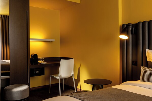 A hotel room with a bed, a desk with a chair, a small round table, and a wall-mounted lamp.