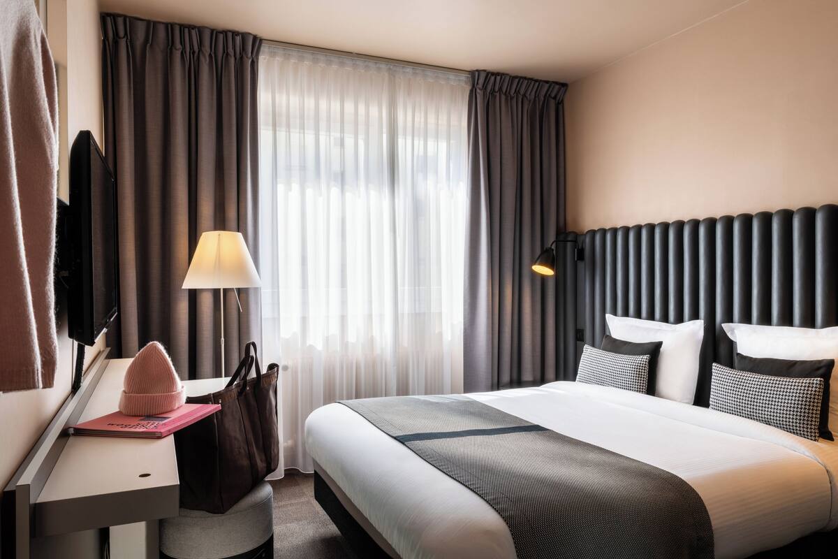 A hotel room with a bed, a desk with a lamp, a television, and a window with curtains.