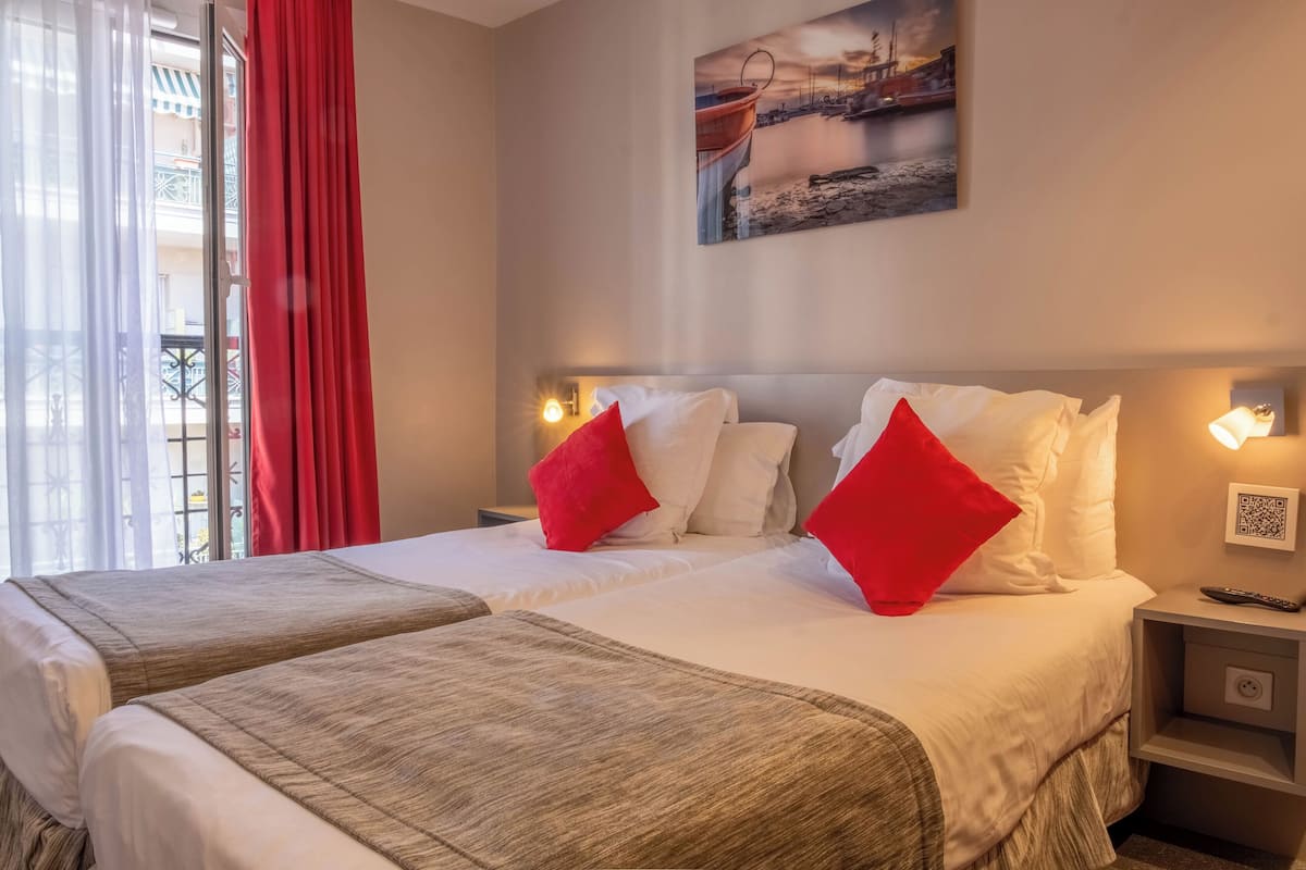 A hotel room with two beds, red and white bedding, a painting on the wall, and a balcony with red curtains.