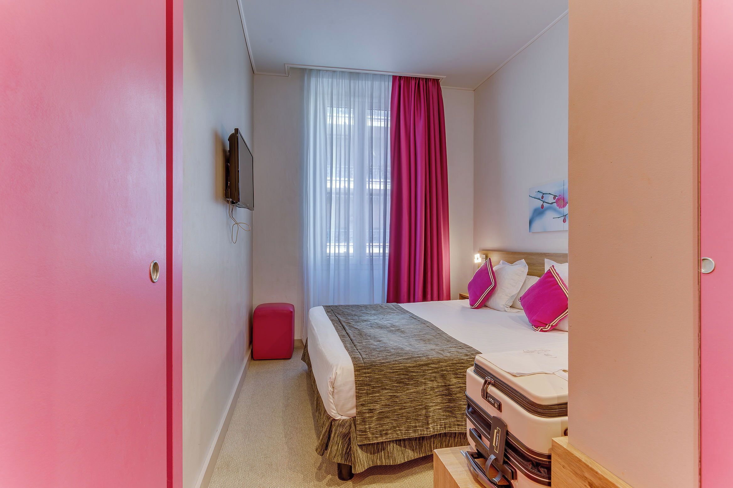 Un dormitori amb un llit, una paret vermella, una finestra amb cortines lleugeres, un coixí rosa i un petit tamboret vermell.