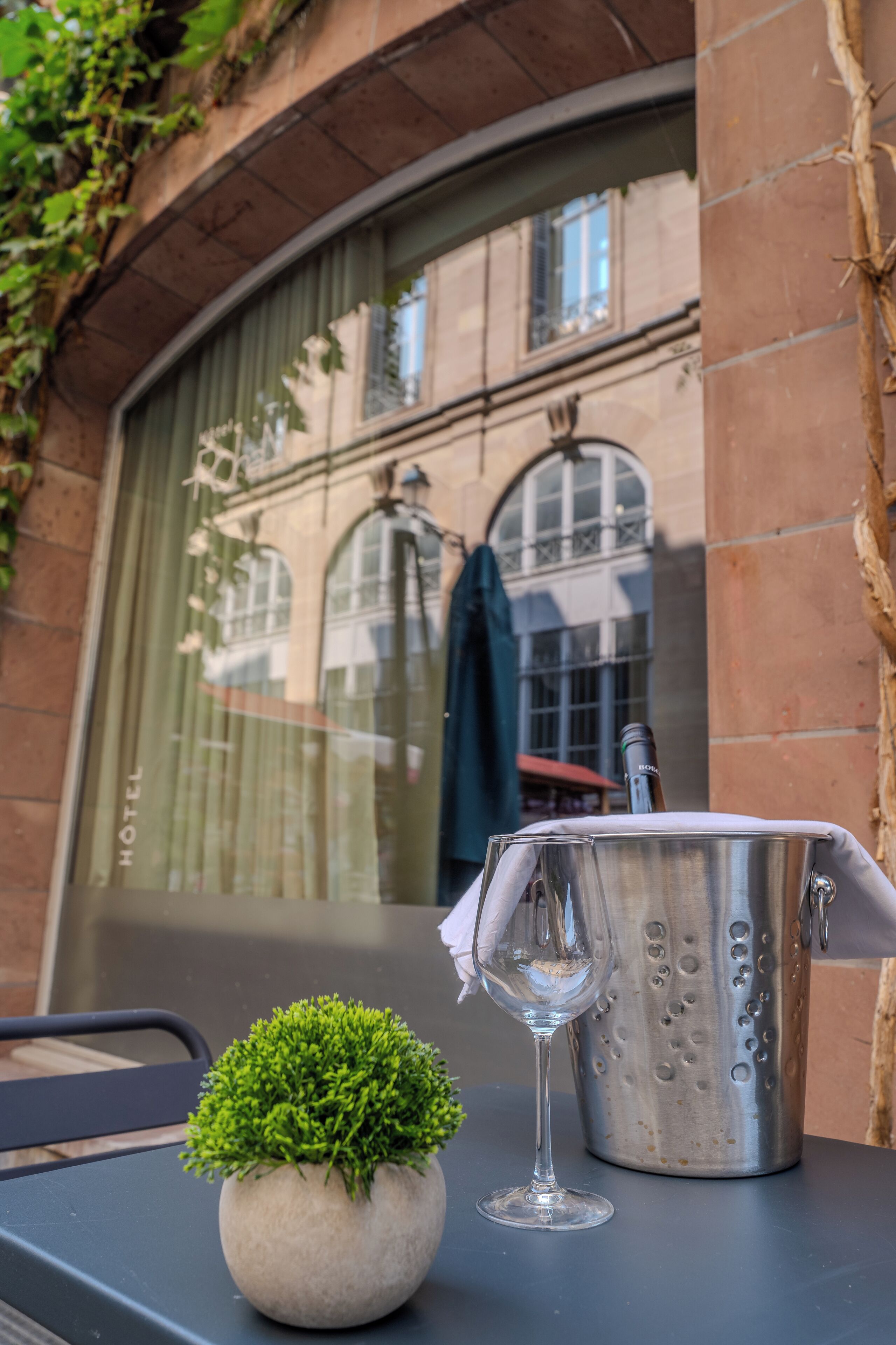Una mesa con una planta en maceta, una copa de vino y un recipiente metálico, probablemente un cubo para champán, frente a un edificio con un amplio ventanal y una puerta.