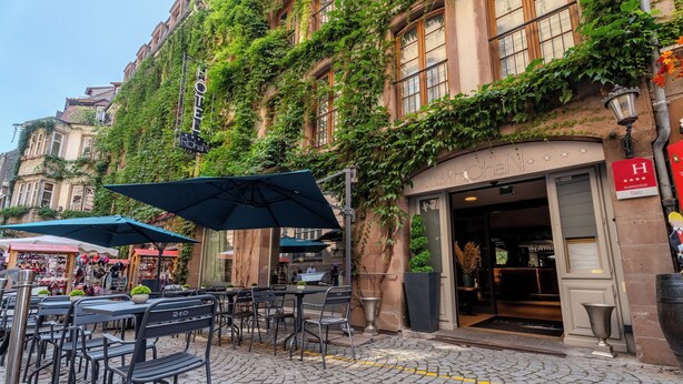 Un café al aire libre con mesas y sillas, adoquinado y un edificio con paredes cubiertas de hiedra.