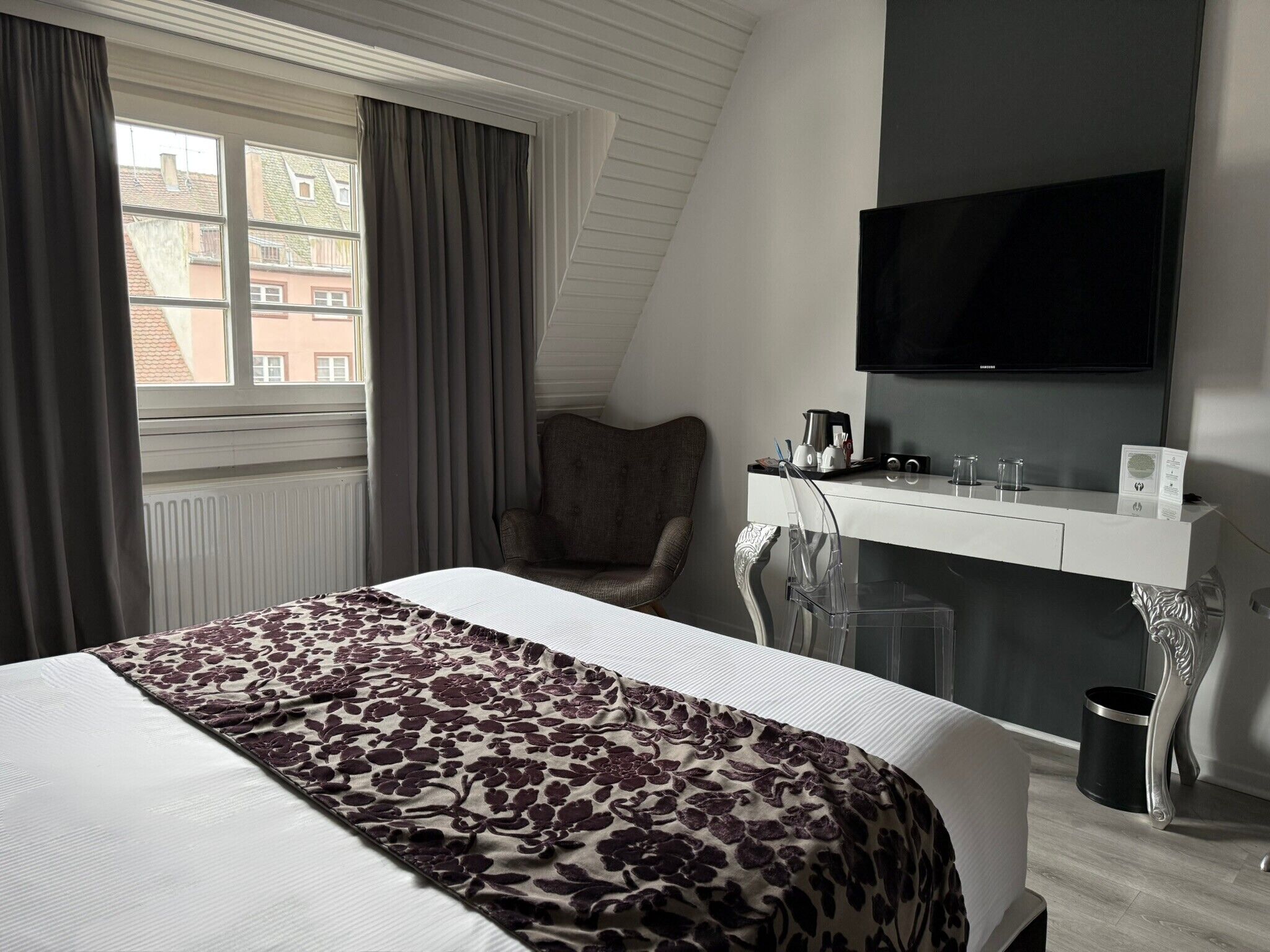 A hotel room with a bed, a desk with a flat-screen TV, a chair, and a window with curtains.