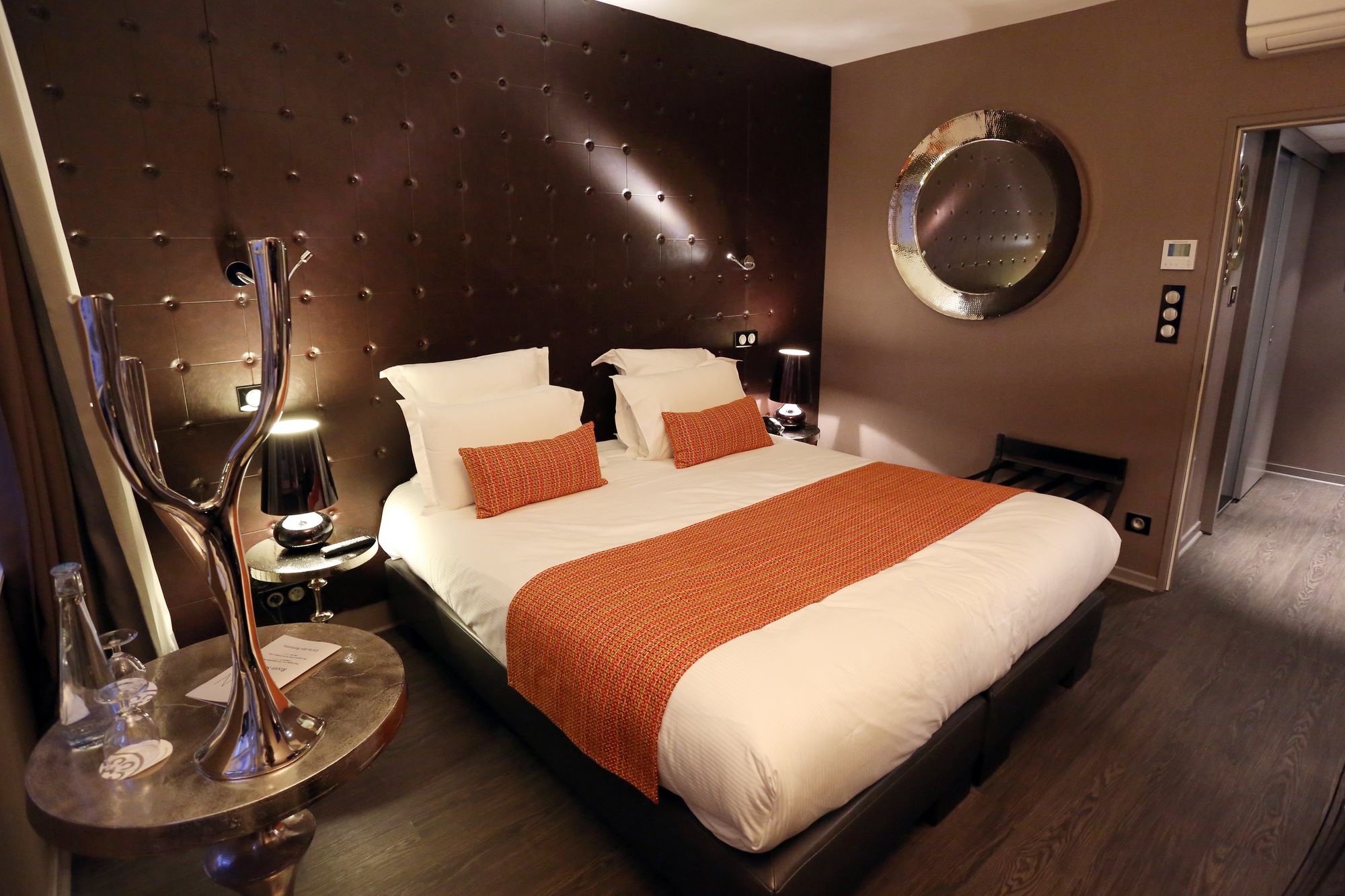 Una habitación de hotel con una cama grande, un espejo redondo, una mesita de noche con lámpara y un calefactor en la pared.