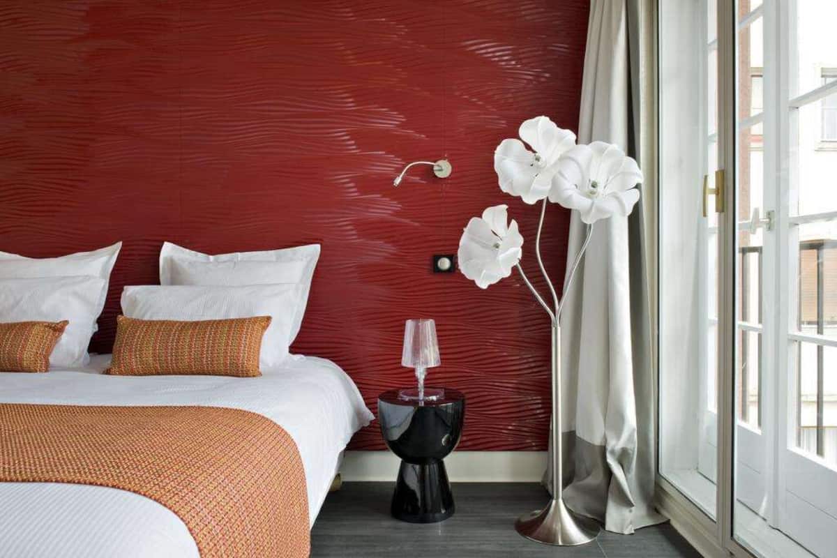 A hotel room with a red textured wall, a bed with white bedding, a black side table, and a white orchid vase.
