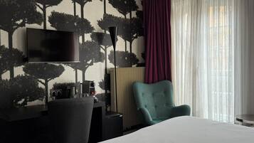 Quarto de hotel moderno com uma cama grande, uma escrivaninha com televisão de tela plana, uma poltrona verde-água e uma janela ampla com cortinas translúcidas.