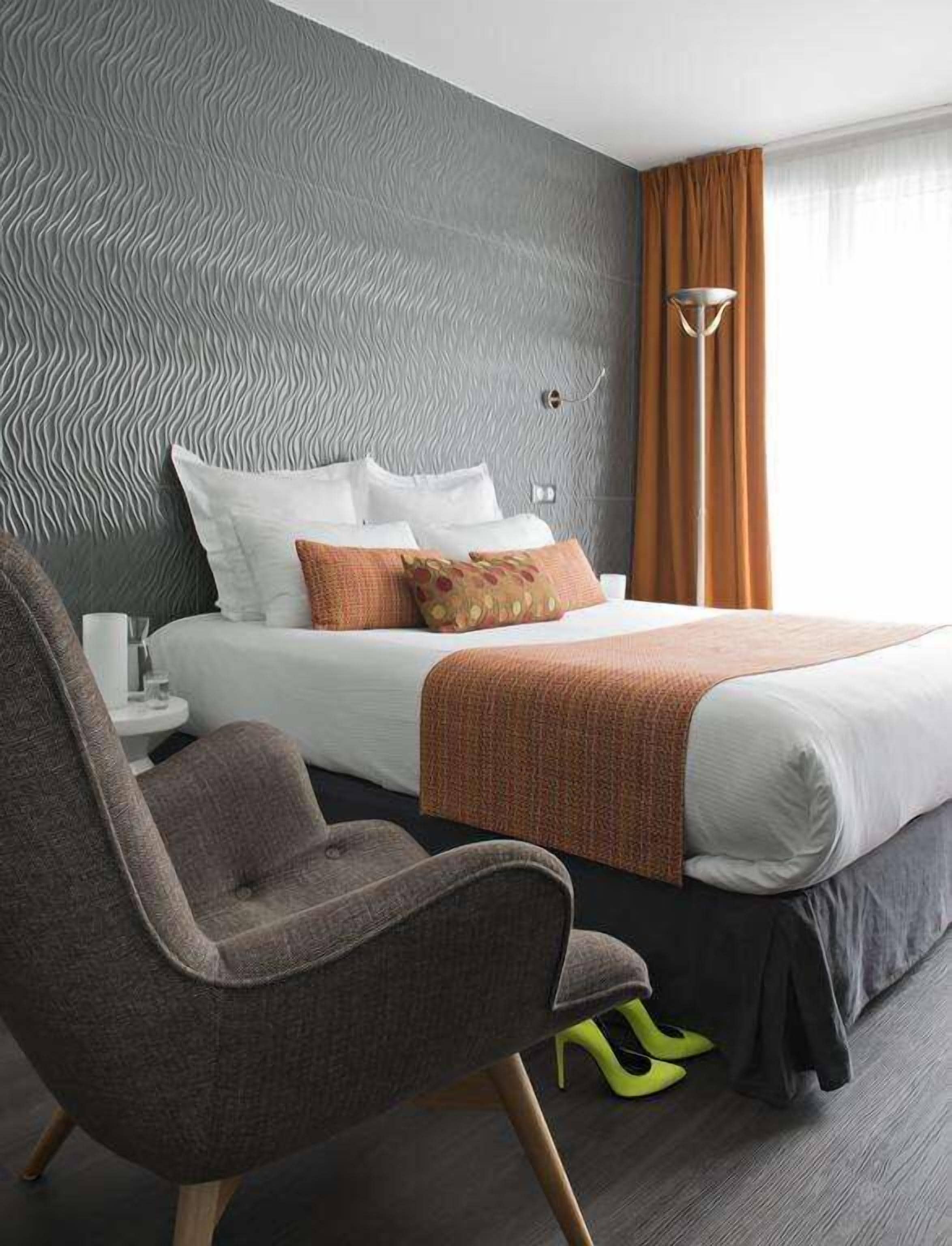 Une chambre d’hôtel moderne avec un mur gris à texture, un lit bien fait avec du linge de lit blanc et des coussins orange, un fauteuil gris et une lampe sur pied.