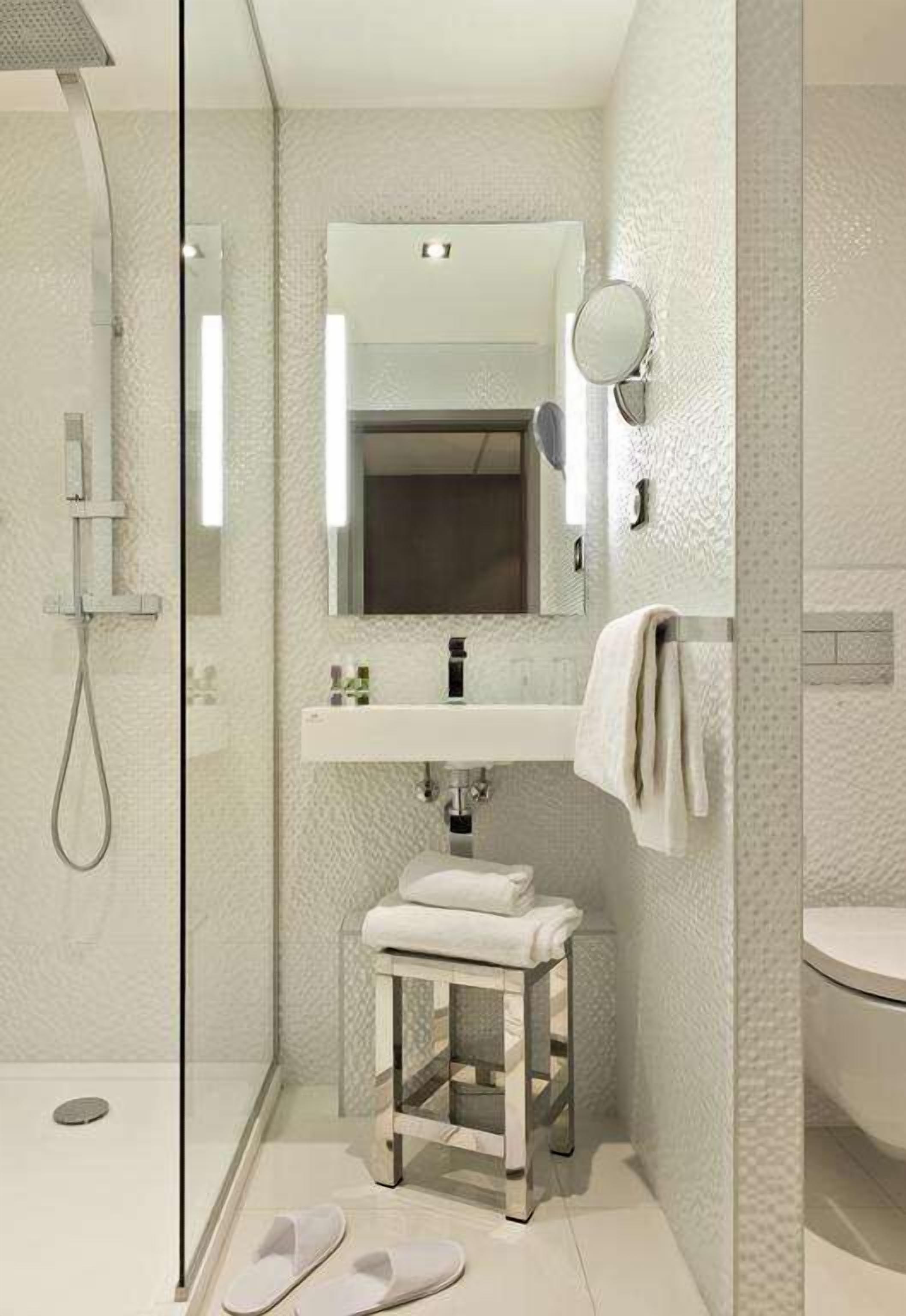 A modern bathroom with a glass shower enclosure, a white sink with a mirror above it, and a small stool.
