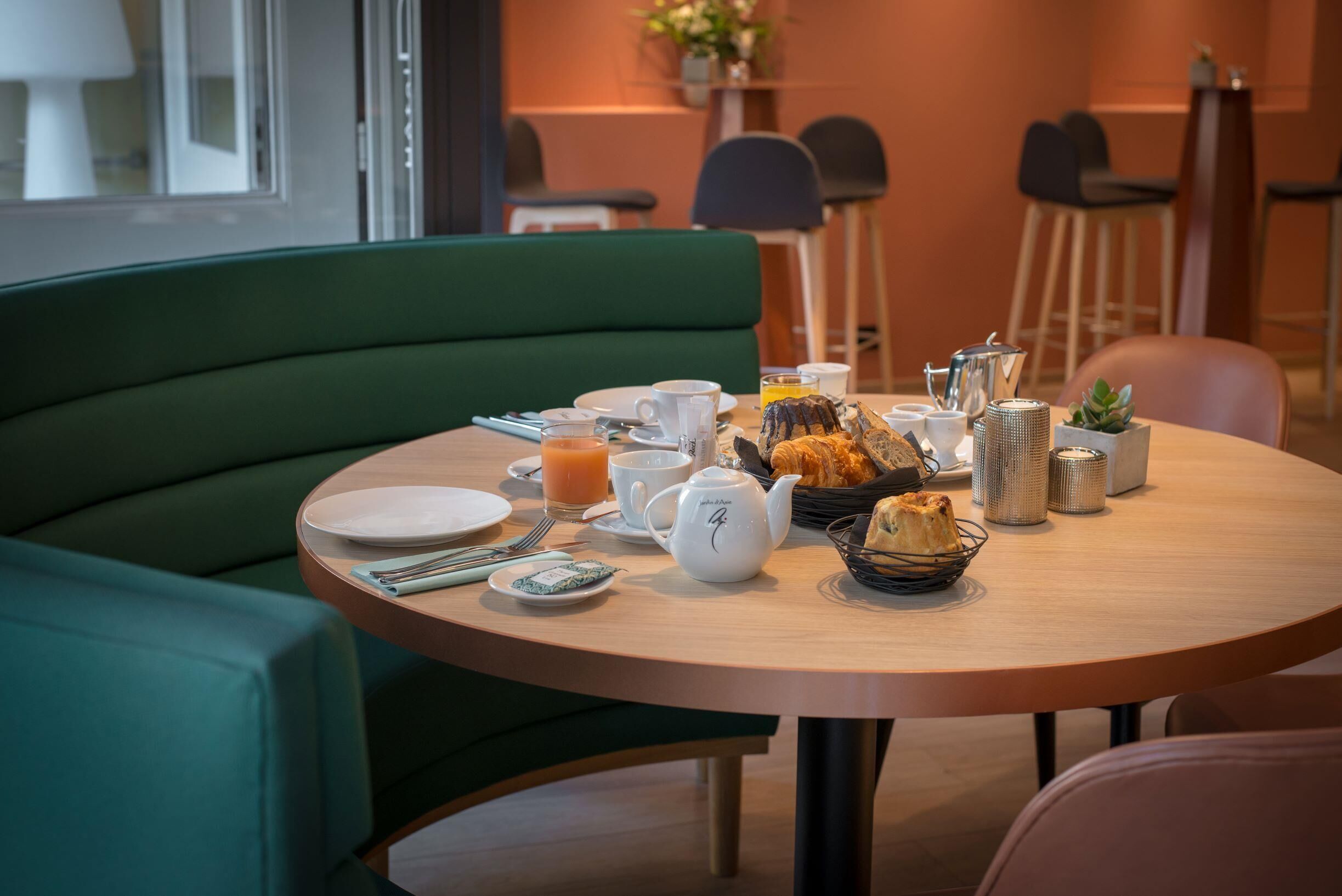 Une table de petit-déjeuner dressée avec des assiettes, des tasses, une théière, un panier de viennoiseries et une petite plante.