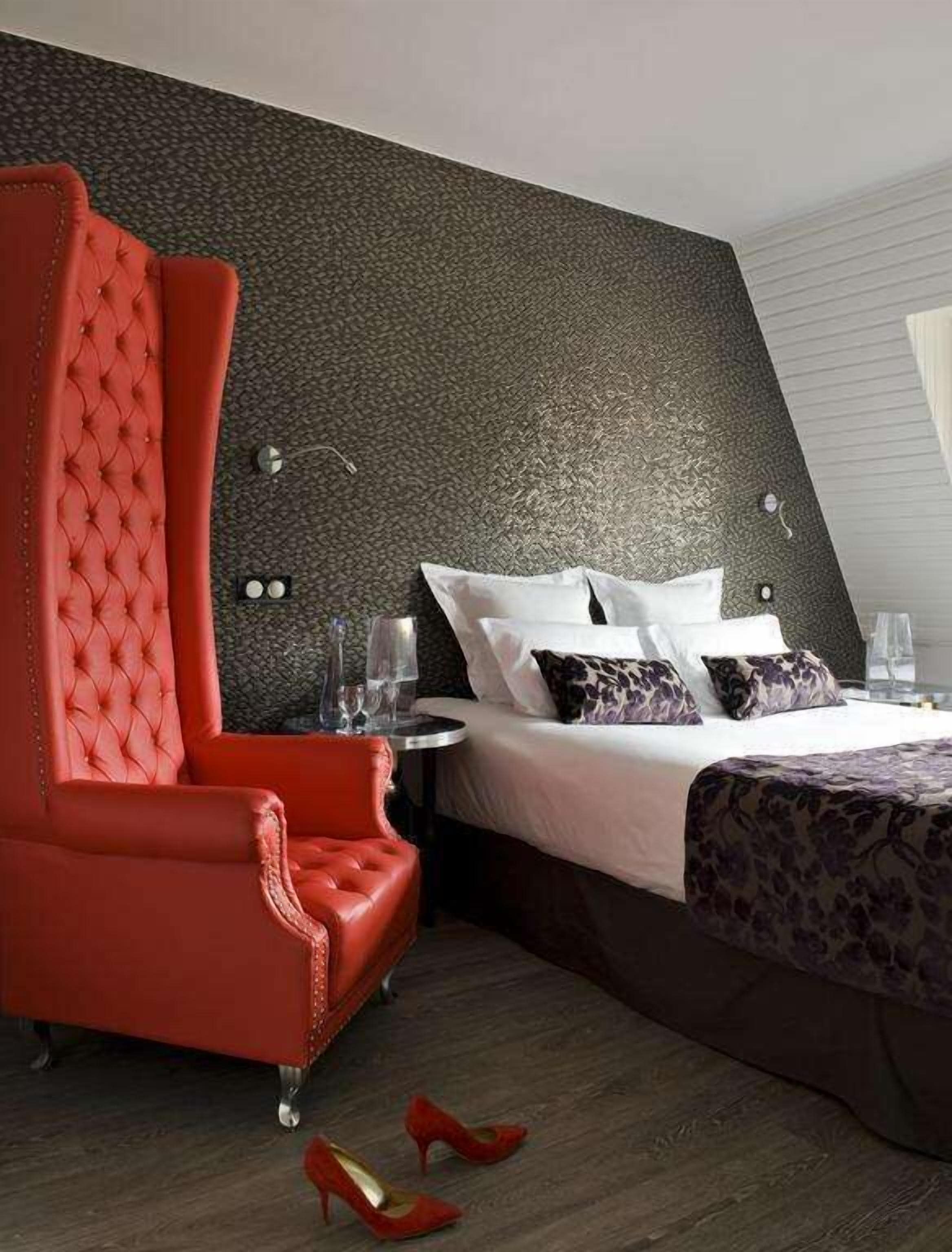 A modern hotel room with a large red armchair, a neatly made bed with white and patterned pillows, and a dark textured wall.