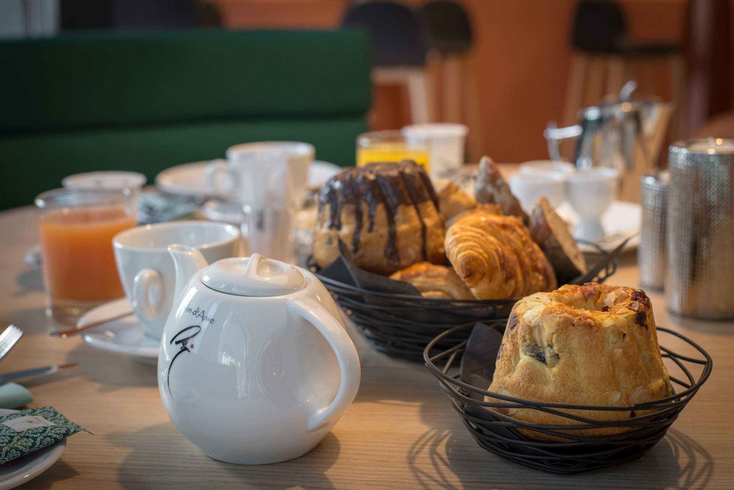 A breakfast table with a teapot, cups, a basket of croissants, and a muffin.