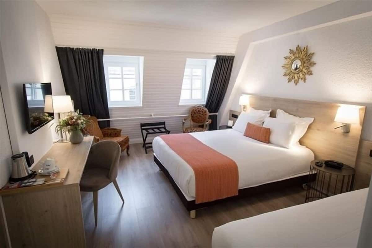 A hotel room with a large bed, a desk, a chair, a small table, and a round chair. There are two windows with curtains, a wall-mounted mirror, and a decorative sunburst light fixture.