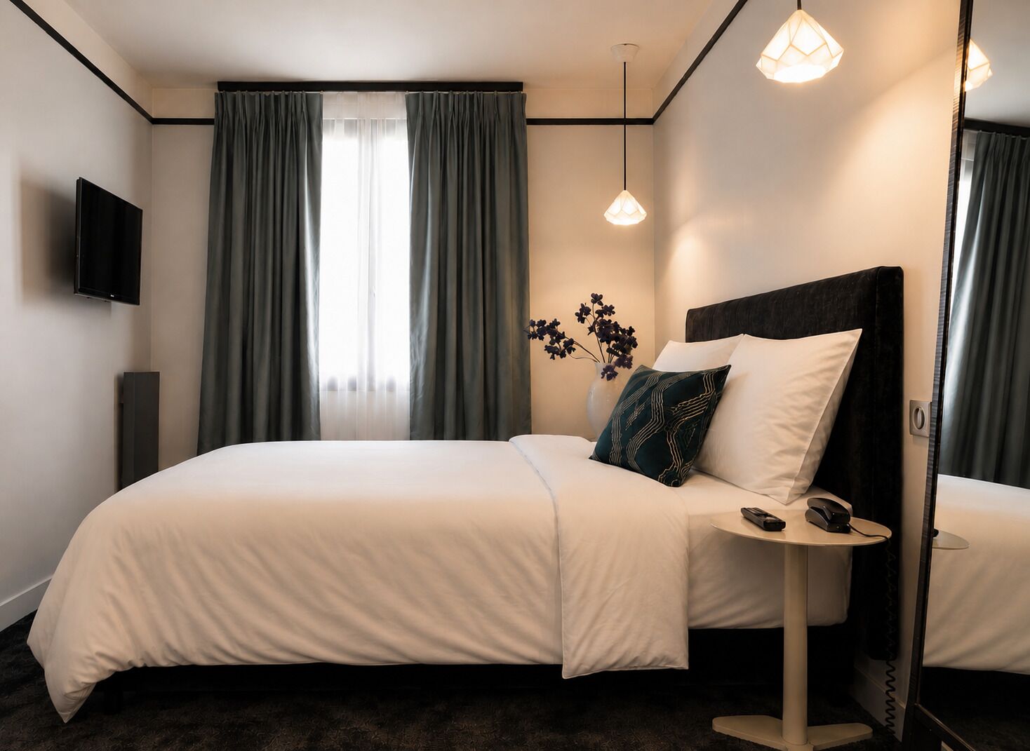 Habitación de hotel con cama, mesita de noche, televisor de pantalla plana, un jarrón con flores y un espejo grande.