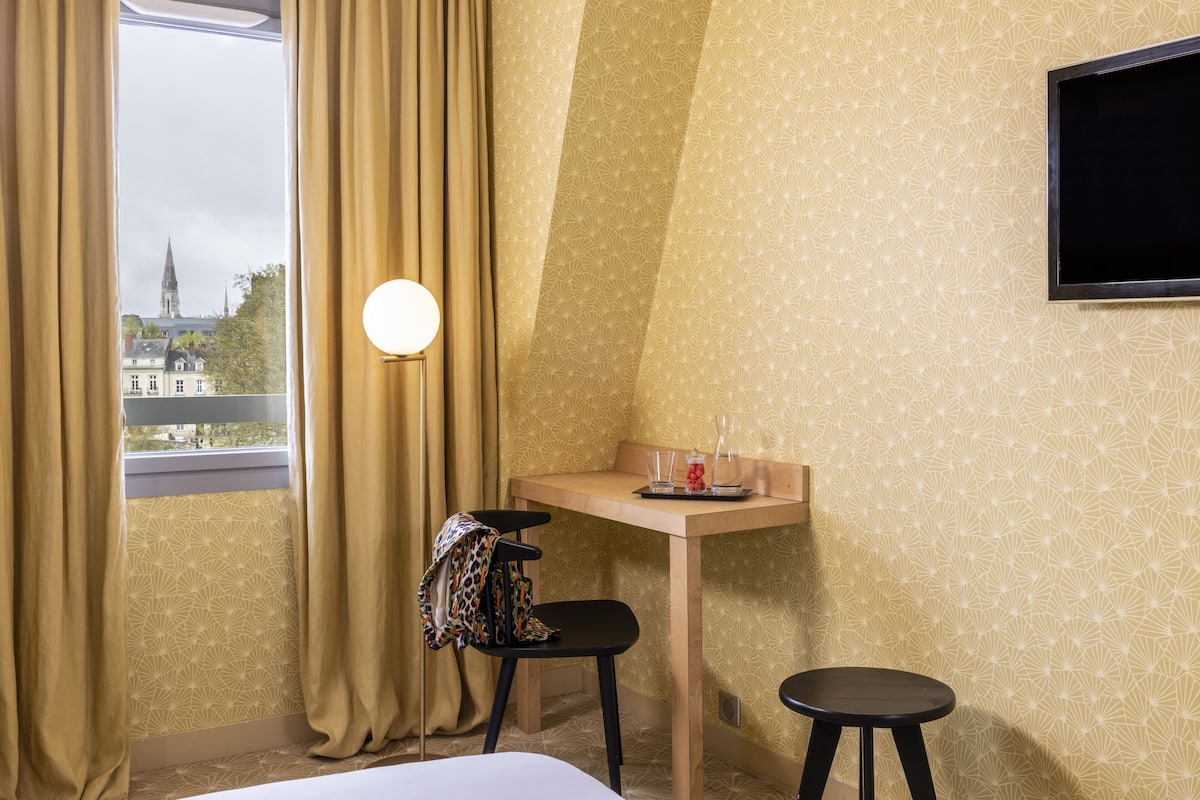 Ein Hotelzimmer mit Bett, Schreibtisch mit Stuhl, Wandfernseher und einem Vorhangfenster.
