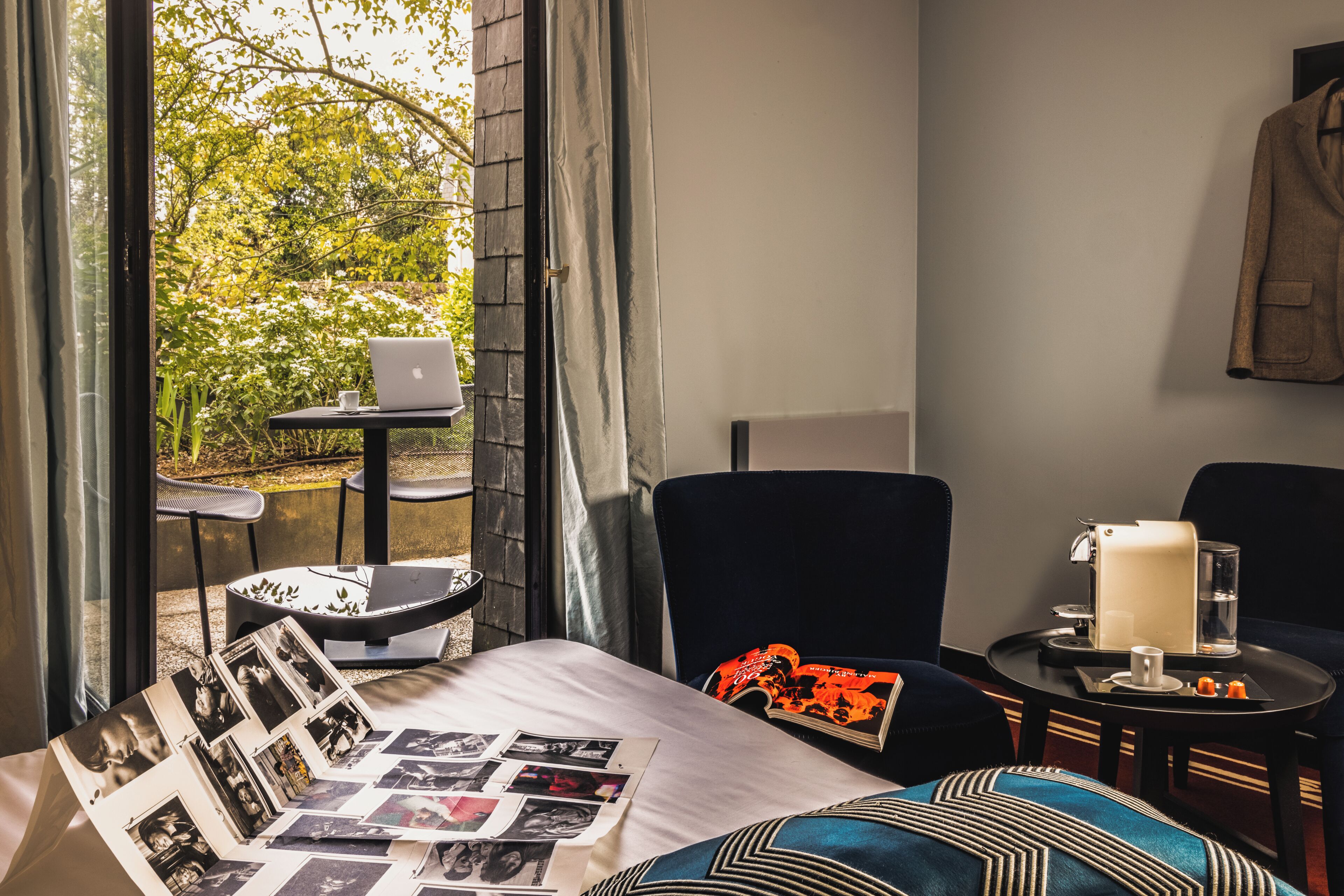 A room with a bed, a laptop on a small table, a chair, a coffee machine, and a view of greenery outside.